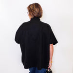 Person wearing a black cape  and blue pants against a white wall