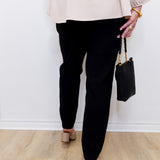 Person wearing black pants and a beige top, holding a black handbag against a white wall.