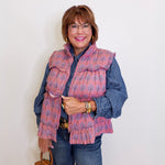 Woman wearing a patterned pink vest over a denim shirt against a white wall.