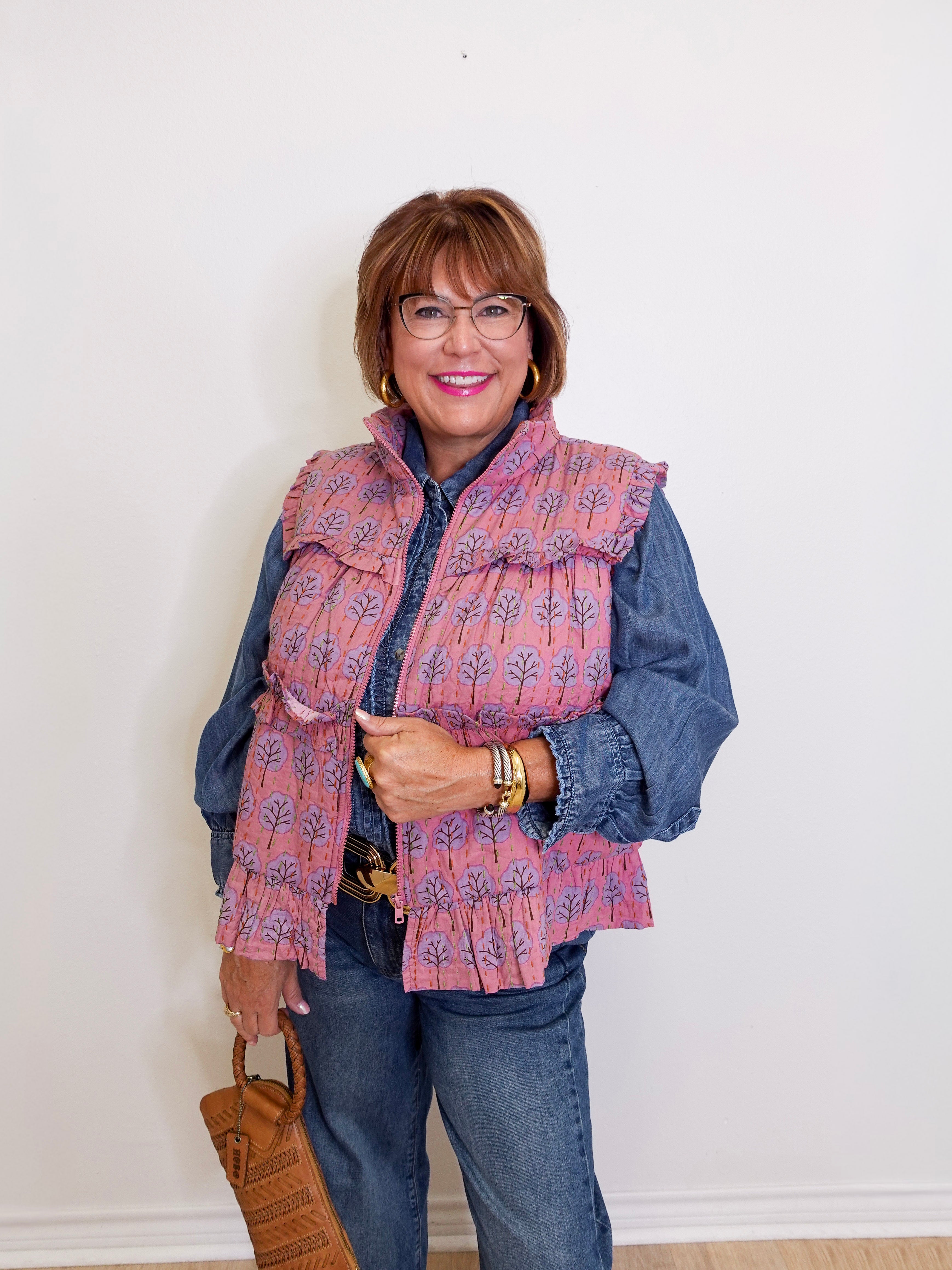Woman wearing a patterned pink vest over a denim shirt against a white wall.