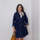 Woman wearing a denim dress with a scarf and holding a bag against a white wall.