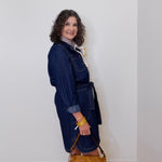 Woman wearing a denim dress holding a brown handbag against a white wall.