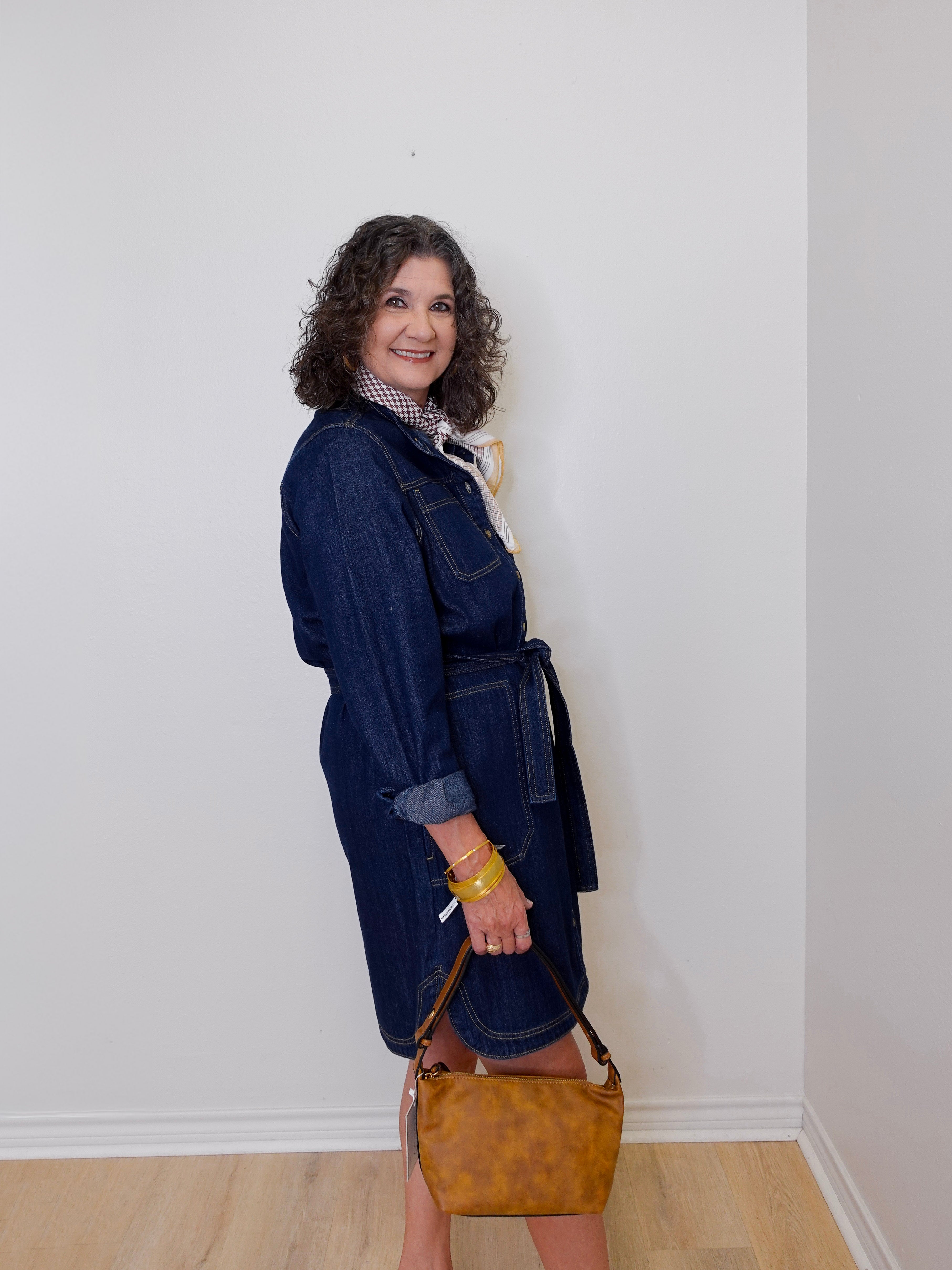 Woman wearing a denim dress holding a brown handbag against a white wall.
