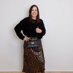 Woman wearing a black top and leopard print skirt holding a black handbag against a white wall.