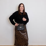 Woman wearing a black top and leopard print skirt holding a black handbag against a white wall.