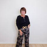 Woman wearing a black top and patterned pants standing against a white wall.