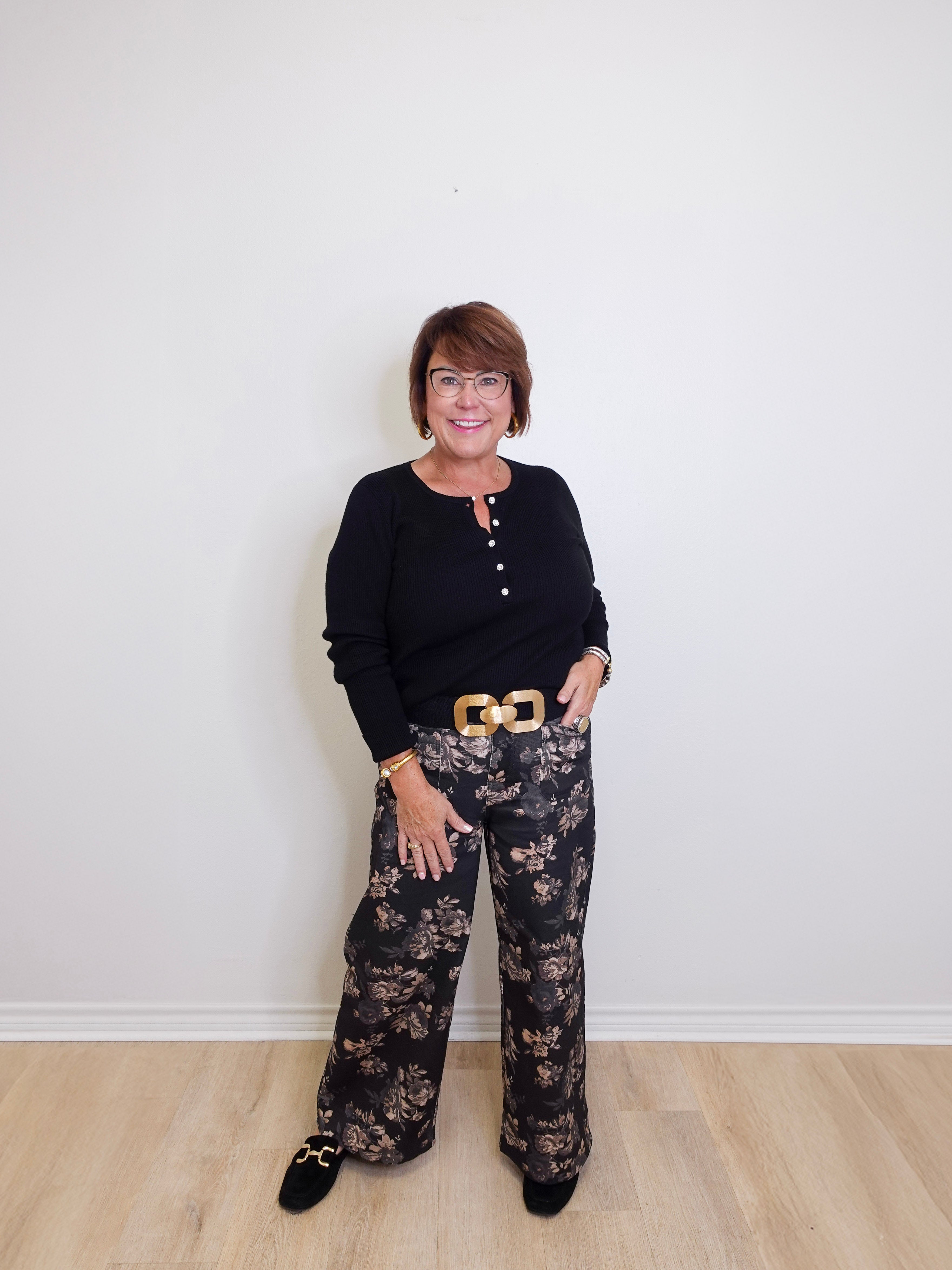 Woman wearing a black top and patterned pants standing against a white wall.