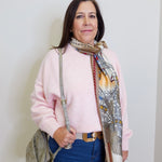 Woman wearing a pink sweater and blue jeans with a patterned scarf, standing against a white wall.