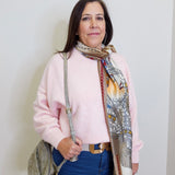 Woman wearing a pink sweater and blue jeans with a patterned scarf, standing against a white wall.