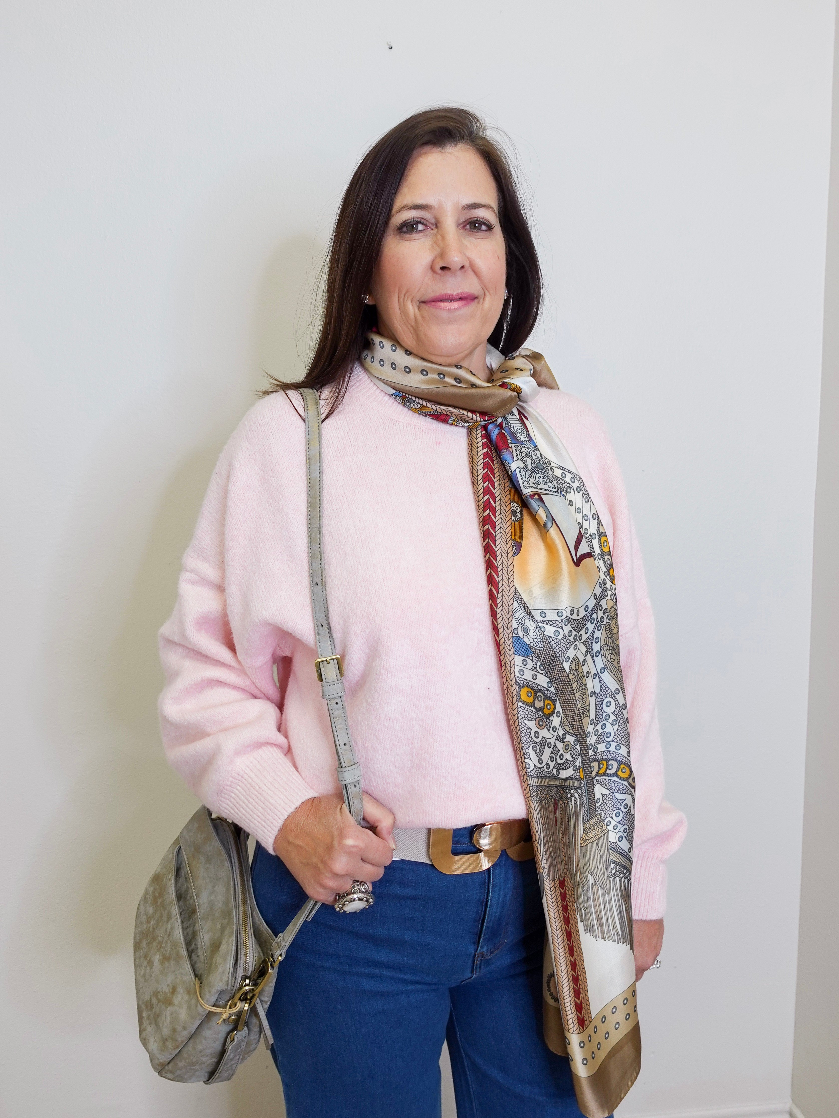 Woman wearing a pink sweater and blue jeans with a patterned scarf, standing against a white wall.