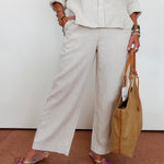 Person wearing beige outfit with wide-leg pants and holding a woven bag on a plain background