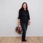 Woman in black polka dot shirt and gray pants holding a colorful bag against a white wall.