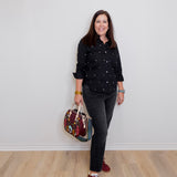 Woman in black polka dot shirt and gray pants holding a colorful bag against a white wall.