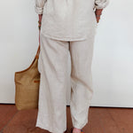 Person wearing a beige outfit with wide-leg pants and holding a brown bag against a white wall.