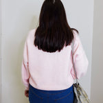 Person wearing a pink sweater and blue jeans standing against a white wall.