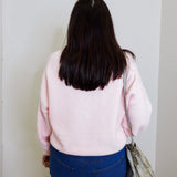 Person wearing a pink sweater and blue jeans standing against a white wall.
