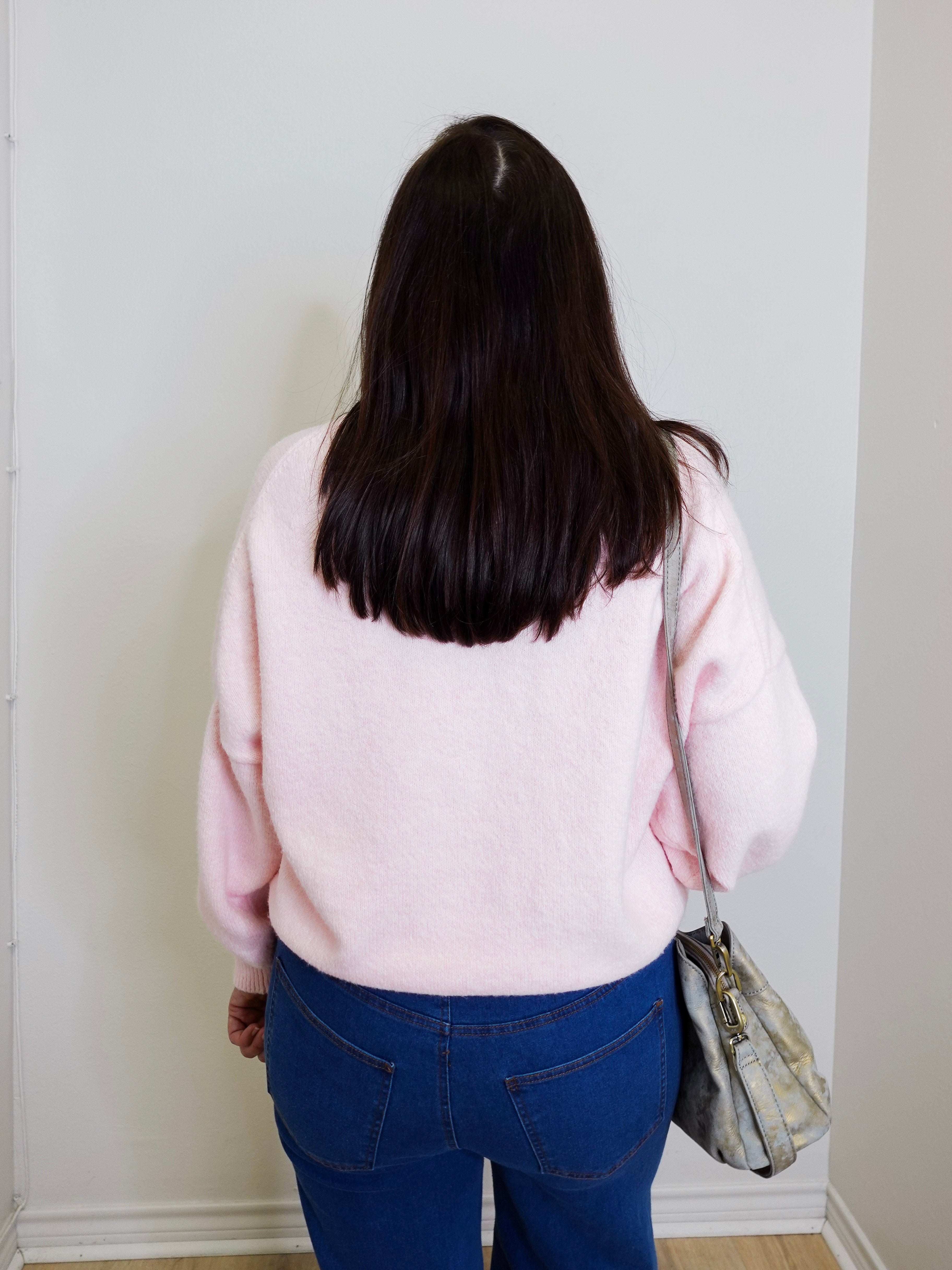 Person wearing a pink sweater and blue jeans standing against a white wall.