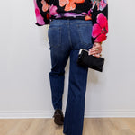 Person wearing a floral blouse and blue jeans holding a black handbag against a white wall.