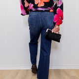 Person wearing a floral blouse and blue jeans holding a black handbag against a white wall.