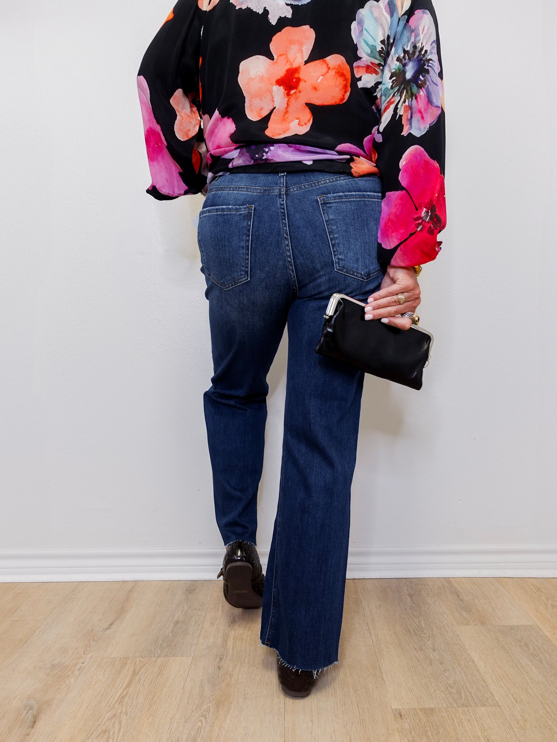 Person wearing a floral blouse and blue jeans holding a black handbag against a white wall.