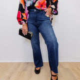 Person wearing a colorful floral blouse, blue jeans, and black shoes on a plain background