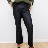 Person wearing a green shirt, black pants, and black loafers with gold accents against a white wall.