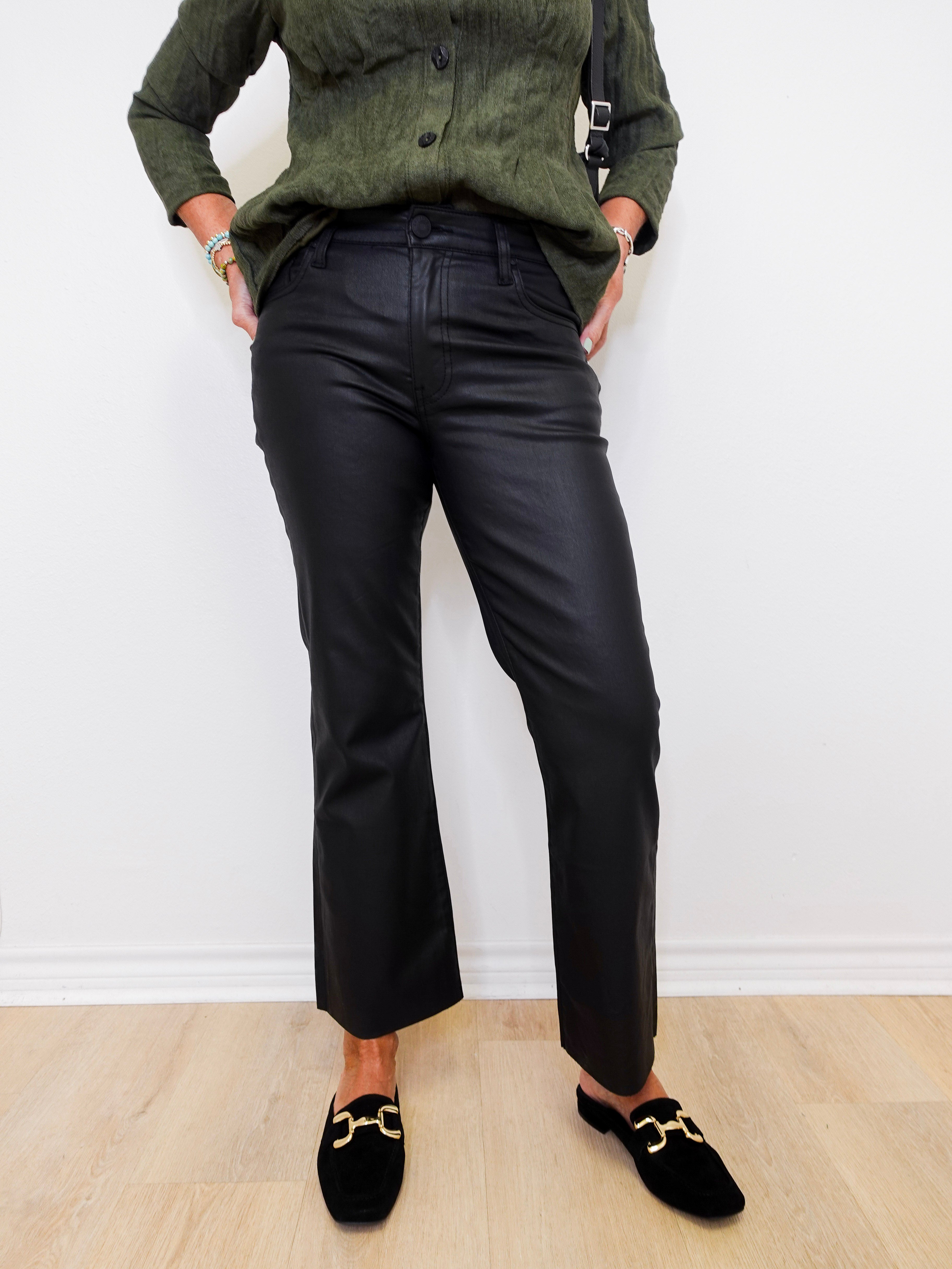 Person wearing a green shirt, black pants, and black loafers with gold accents against a white wall.