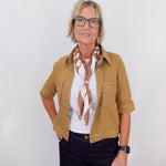 Woman wearing a tan jacket, white shirt, and black pants with a scarf against a white background