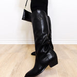 Black knee-high boots on a light wooden floor with a white wall background