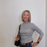 Woman wearing a gray patterned turtleneck and dark jeans against a white wall.