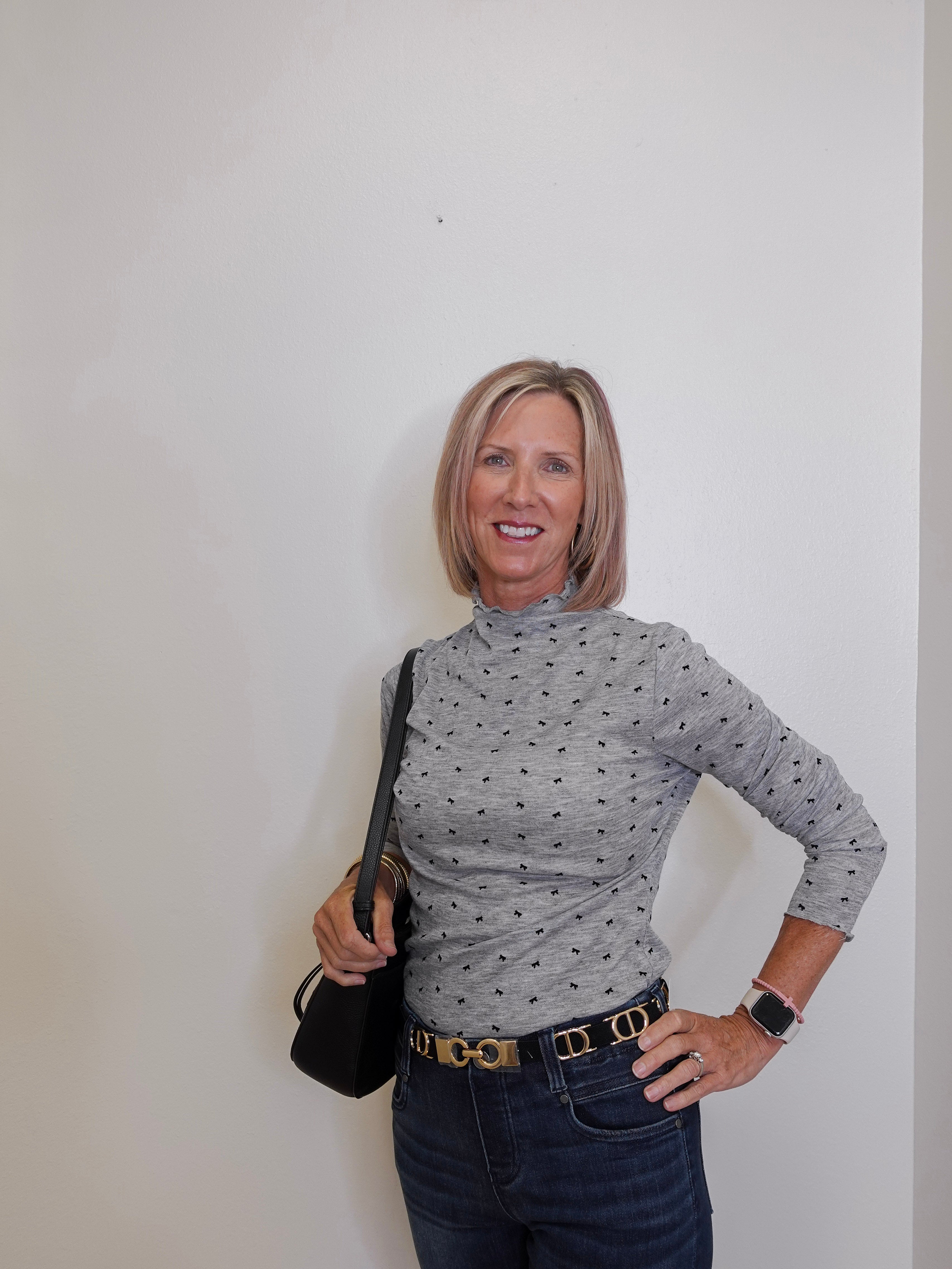 Woman wearing a gray patterned turtleneck and dark jeans against a white wall.