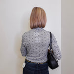Person wearing a gray patterned shirt and dark jeans, standing against a plain white wall.
