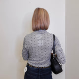 Person wearing a gray patterned shirt and dark jeans, standing against a plain white wall.