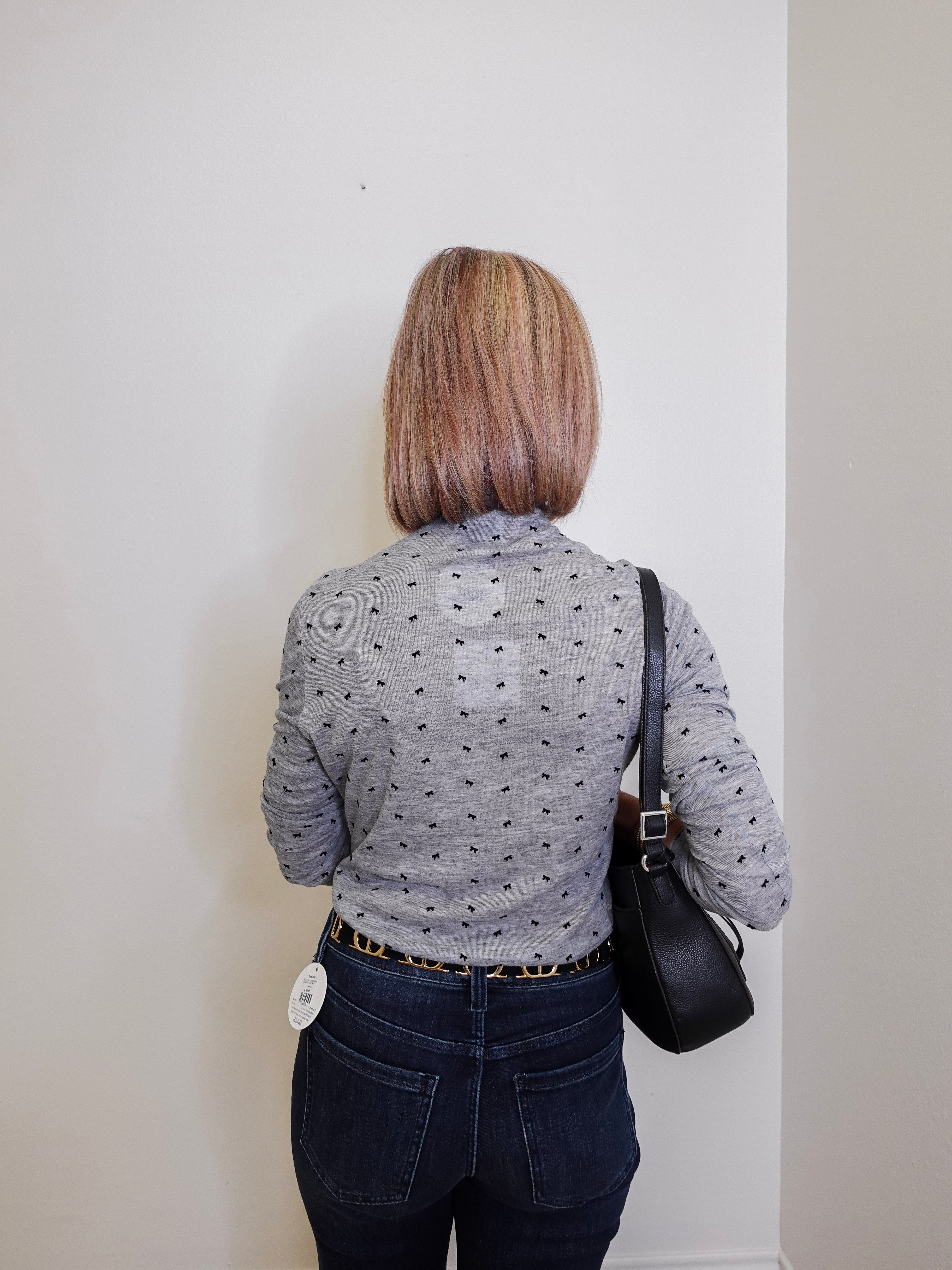 Person wearing a gray patterned shirt and dark jeans, standing against a plain white wall.