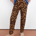 A person wearing leopard-print pants with a blue denim shirt and beige wedge sandals, standing against a white wall.