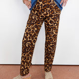 A person wearing leopard-print pants with a blue denim shirt and beige wedge sandals, standing against a white wall.