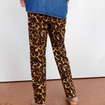 A person shown from the back wearing leopard-print pants, a blue denim top, and wedge sandals against a plain white wall and terracotta floor.