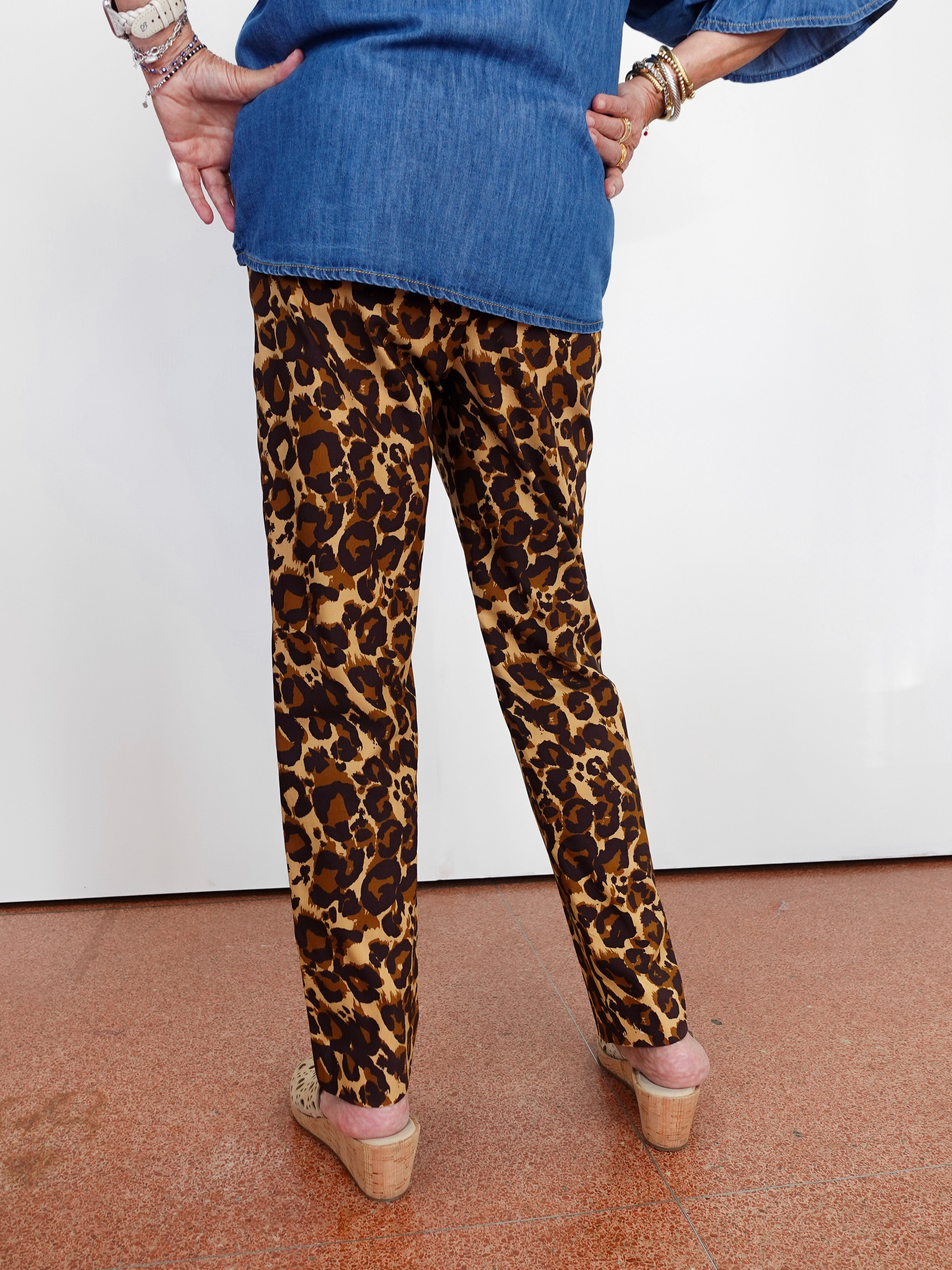 A person shown from the back wearing leopard-print pants, a blue denim top, and wedge sandals against a plain white wall and terracotta floor.