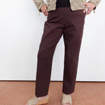 A person wearing dark brown straight-leg trousers, a textured beige and brown jacket, a black top, and woven wedge sandals, standing against a white wall.
