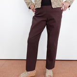 A person wearing dark brown straight-leg trousers, a textured beige and brown jacket, a black top, and woven wedge sandals, standing against a white wall.