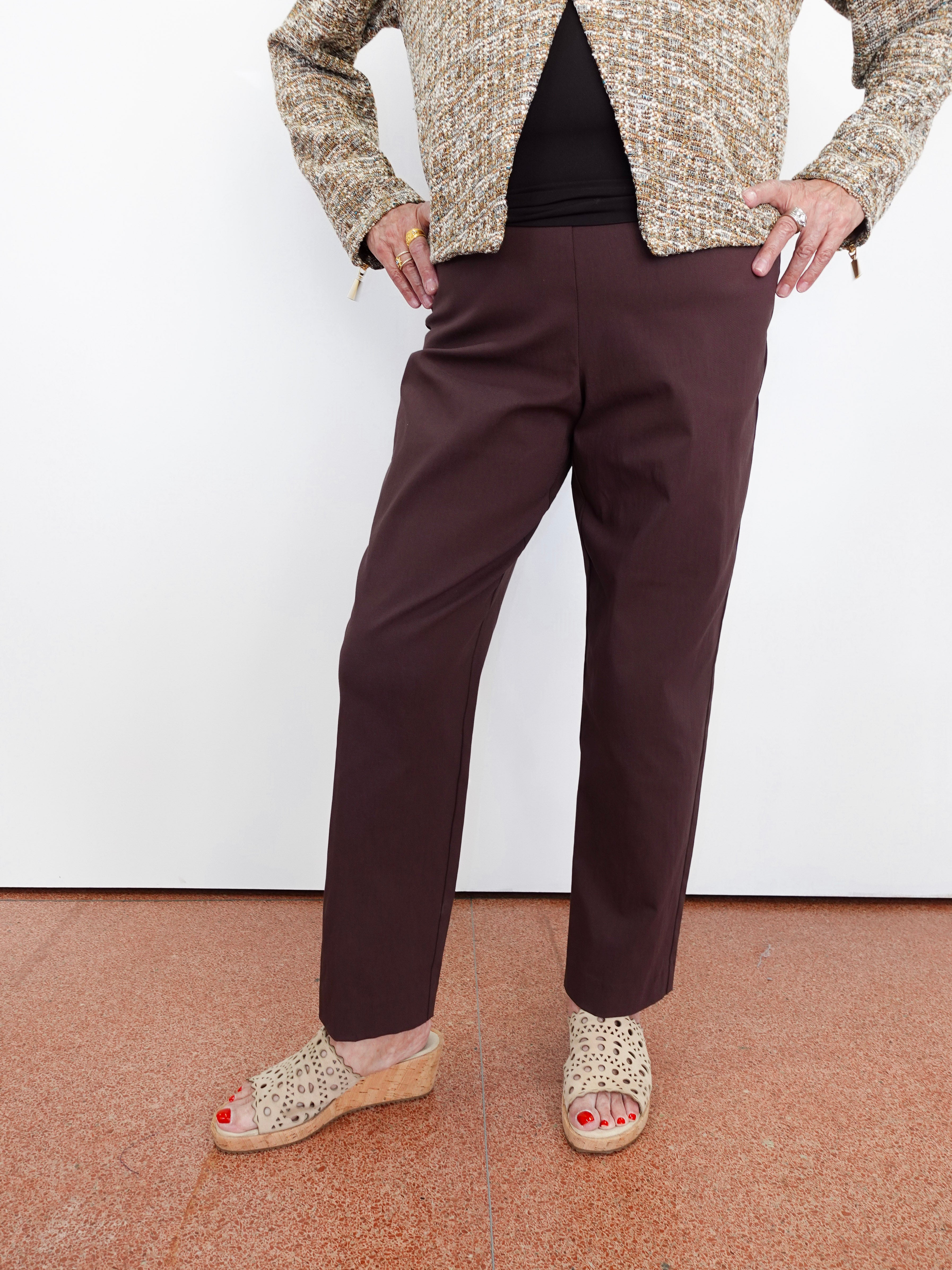 A person wearing dark brown straight-leg trousers, a textured beige and brown jacket, a black top, and woven wedge sandals, standing against a white wall.