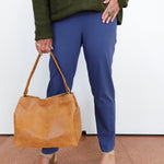 A person wearing blue pants and an olive green sweater holds a large tan leather shoulder bag, standing against a white wall.
