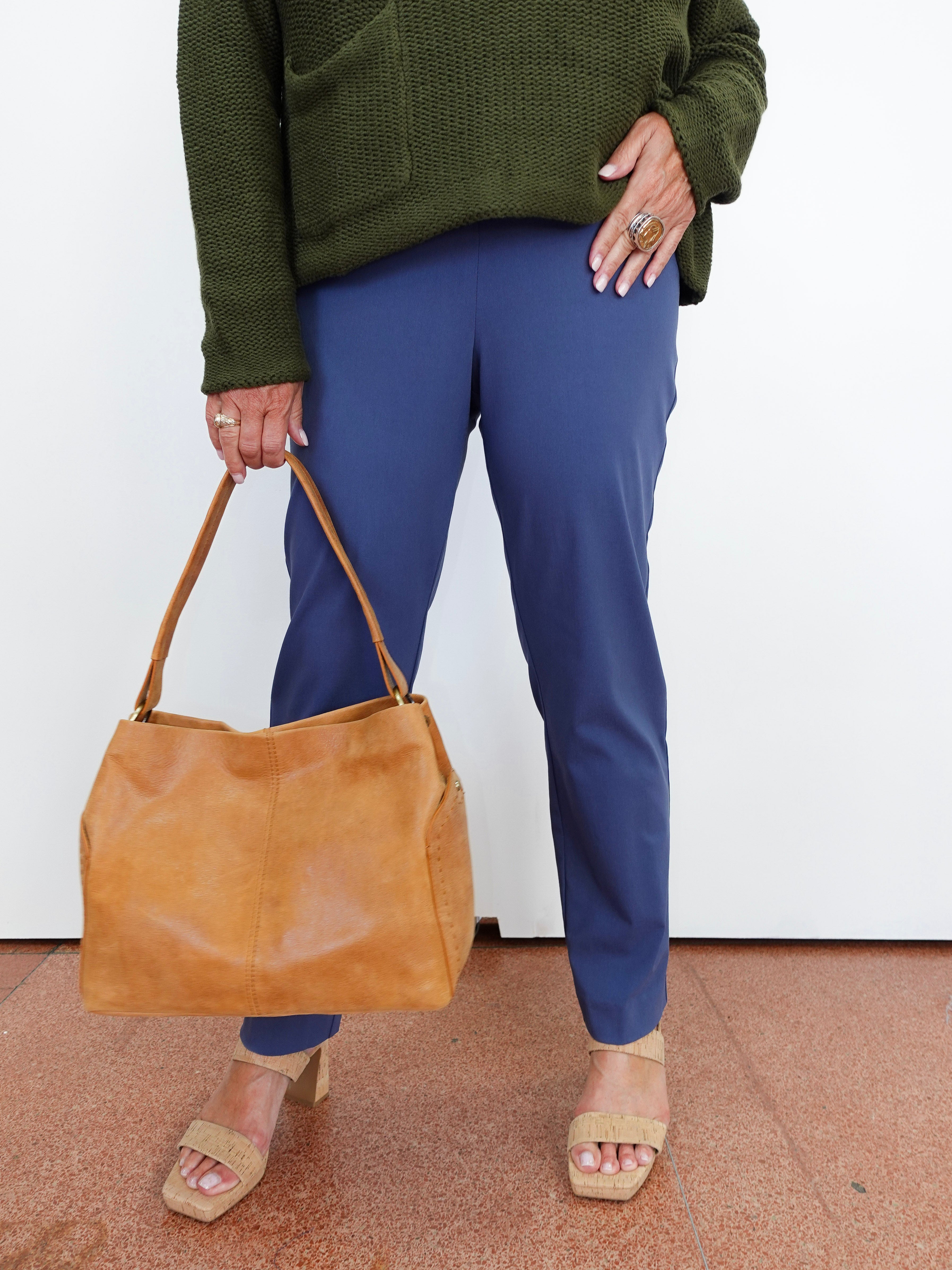 A person wearing blue pants and an olive green sweater holds a large tan leather shoulder bag, standing against a white wall.