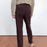 A person shown from the back wearing dark brown straight-leg trousers, a textured beige and brown knit sweater, and wedge sandals, standing against a white wall on a terracotta tiled floor.