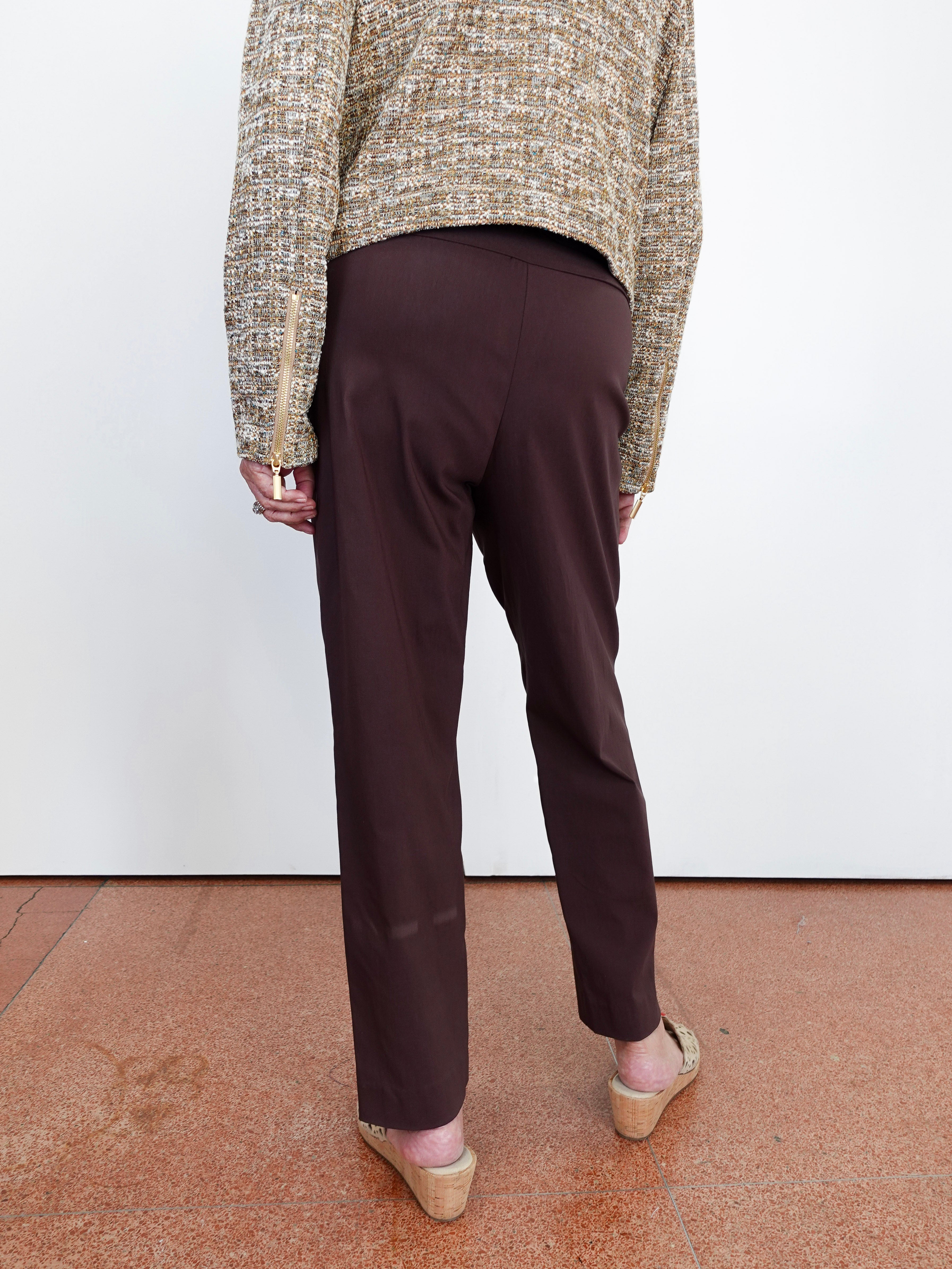 A person shown from the back wearing dark brown straight-leg trousers, a textured beige and brown knit sweater, and wedge sandals, standing against a white wall on a terracotta tiled floor.