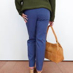 A person shown from the back wearing blue trousers, an olive green sweater, and beige heels, holding a tan leather handbag against a plain white wall.