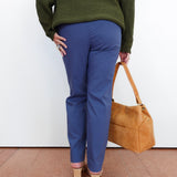 A person shown from the back wearing blue trousers, an olive green sweater, and beige heels, holding a tan leather handbag against a plain white wall.