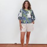 Woman wearing a floral blouse and beige shorts against a white wall.