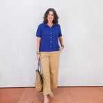 Woman wearing a blue shirt and beige pants standing against a white wall.