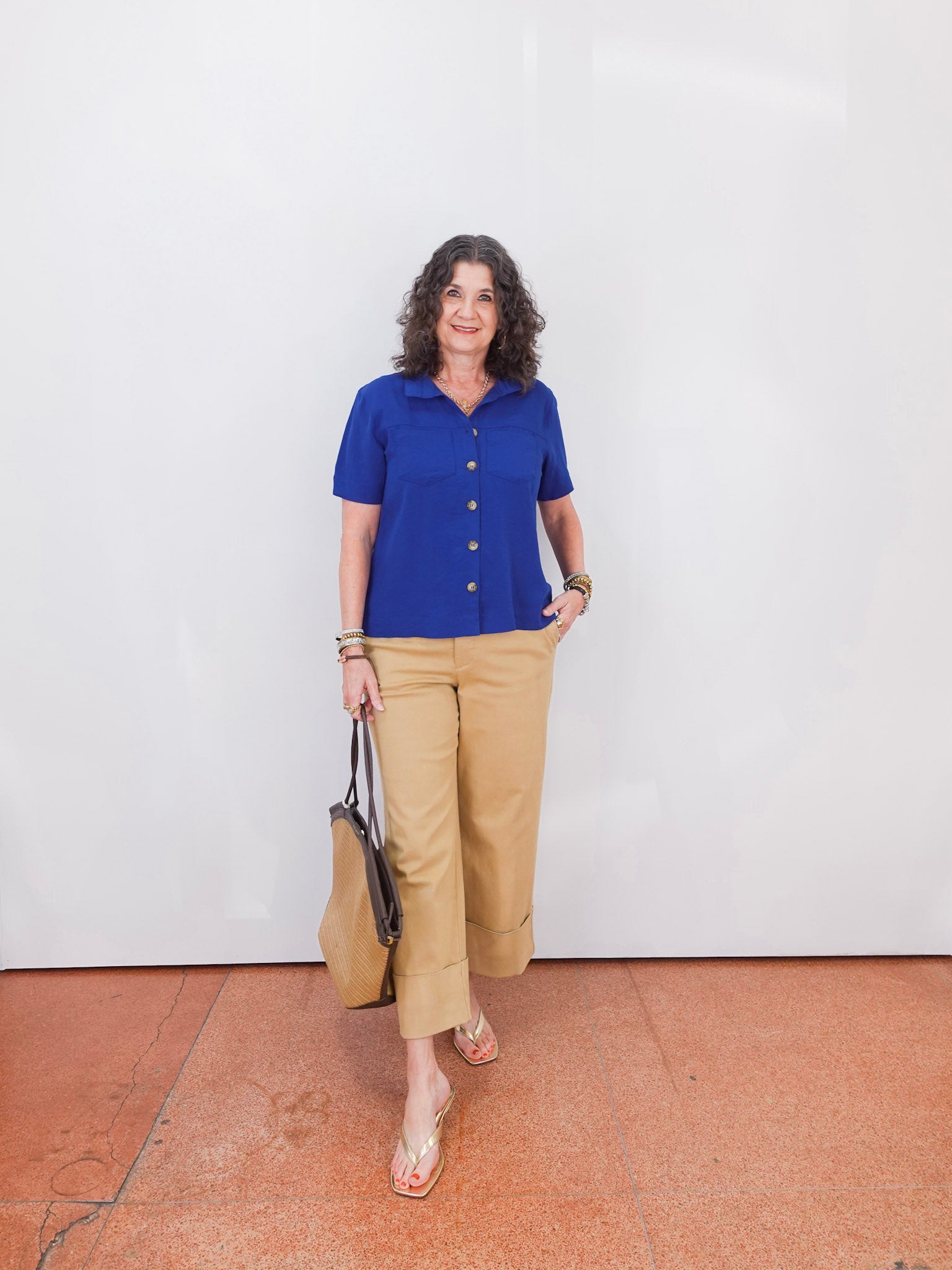 Woman wearing a blue shirt and beige pants standing against a white wall.